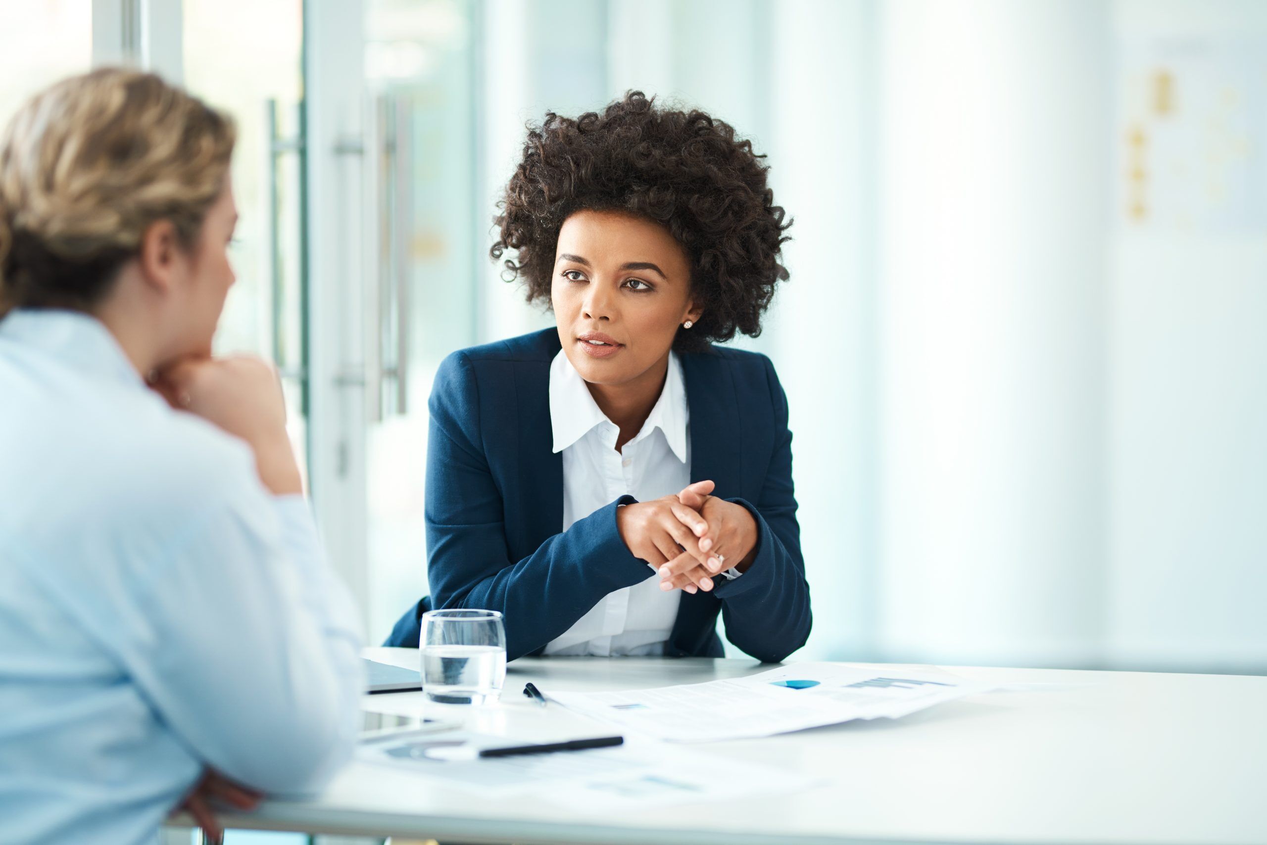 A photo of two business women seated at a table and talking while one is providing input to the other